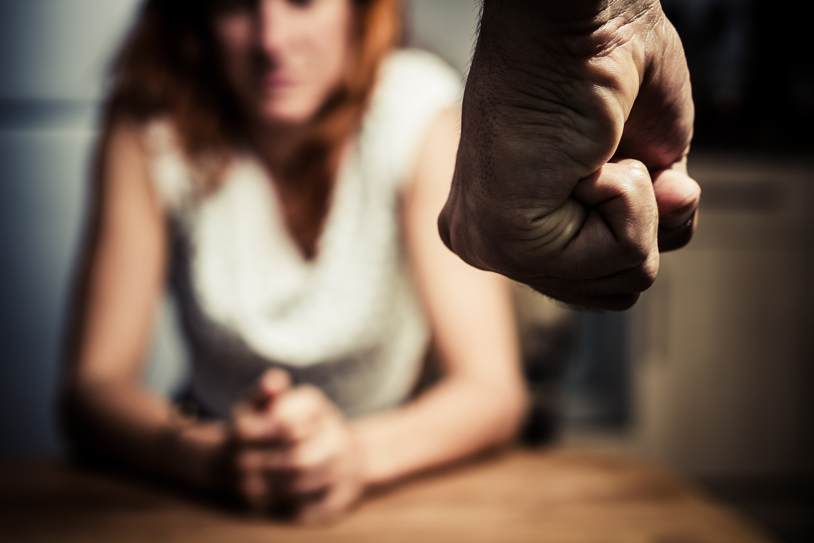 Young woman is sitting hunched at a table at home, the focus is on a man's fist in the foregound of the image