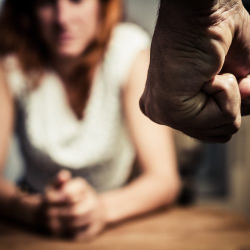 Young woman is sitting hunched at a table at home, the focus is on a man's fist in the foregound of the image